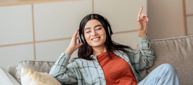 Kanepede oturan genç bir kadın, kulaklık takıyor ve müzik dinlerken gülümsüyor. Rahat ve mutlu görünüyor, rahat bir yaşam alanında bir anlık sevinç gösteriyor..