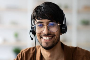 Gözlüklü ve kulaklıklı genç bir adam ev ofisinde konuşurken gülümsüyor. Parlak ışıklandırma ve minimal dekor uzak çalışma için hoş bir atmosfer yaratıyor.