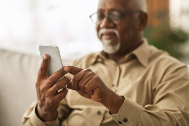 Gözlüklü yaşlı bir adam evdeki bir kanepede oturuyor, akıllı telefonuyla meşgul. Cihazla etkileşime girerken odaklanmış ve memnun görünüyor. Etrafı rahat bir ortamla çevrili..
