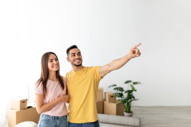 Genç bir çift, karton kutularla dolu yeni evlerinde birlikte duruyorlar. Gülümsüyorlar, kadın sevinçle bakarken erkek heyecan verici bir şekilde çerçeveden bir şeyi işaret ediyor..