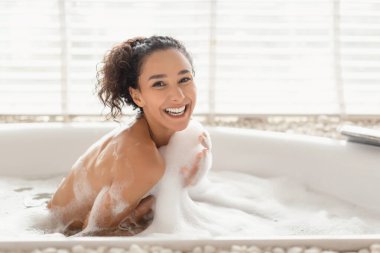 Bir kadın köpük banyosunda eğleniyor, ferah bir küvette rahatlarken ışıl ışıl gülümsüyor..