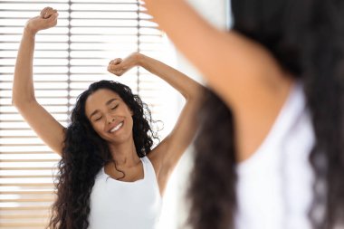 Kıvırcık saçlı bir kadın aynanın önünde kollarını başının üzerine uzatıyor. Yumuşak ışık, ahşap panjurların arasından süzülüp sıcak bir atmosfer yaratır..