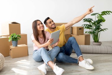Genç bir çift yeni evlerinde karton kutularla çevrili yerde oturuyor. Gülümsüyorlar ve tabletteki bir şeye işaret ediyorlar, heyecanlı bir anı paylaşıyorlar..