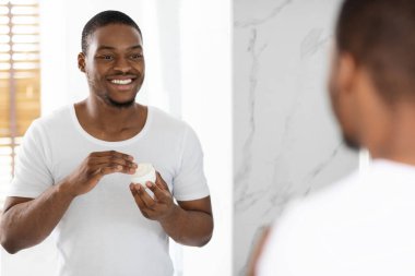 Gülümseyen bir adam nemlendirici bir cilt bakımı uygularken banyo aynasının önünde duruyor. Parlak doğal ışık odayı dolduruyor, neşeli bir atmosfer yaratıyor..