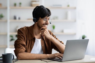 Genç bir adam, parlak ve modern bir ofis bilgisayarına odaklanmış durumda. Kulaklık takıyor ve ahşap bir masada otururken ekranla meşgul olurken gülümsüyor..