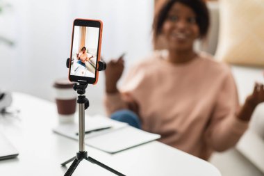 Bir kadın aktif olarak bir video çağrısına katılıyor, masada otururken gülümsüyor ve el kol hareketi yapıyor. Sahne, modern dekorlu ve yakınlarda bir kahve fincanı olan rahat bir ortam yakalıyor..