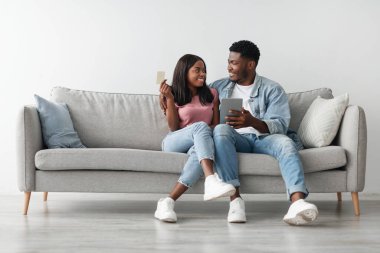 Ticaret ve Kolay Ödeme. Mutlu milenyum siyahi çifti evlerinde kanepede oturup tablet kullanarak ne alacaklarını seçiyorlar ellerinde bankamatik kartı, birbirlerine bakıyorlar, bedava fotokopi çekiyorlar.