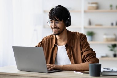 Genç bir adam rahat bir ofiste dizüstü bilgisayarda yazarken gülümsüyor. Kulaklık takıyor, sanal bir toplantıda olduğunu ya da müzik dinlediğini gösteriyor. Doğal ışık odayı dolduruyor..