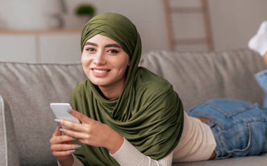 Yeşil başörtüsü takmış genç bir kadın kanepede rahat bir şekilde uzanıyor, akıllı telefonuyla konuşurken sıcak bir şekilde gülümsüyor. Konforlu oda ayarları rahatlatıcı atmosferi arttırır..