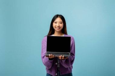 Genç bir kadın, önünde dizüstü bilgisayar tutarak, rahat mor bir kazak giyerek kendine güvenerek ayakta duruyor. Açık mavi arka plan neşeli ve davetkar bir atmosfer yaratır..