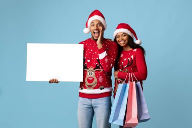 Neşeli bir çift açık mavi arka plana karşı poz veriyor, her biri Noel süveteri giyiyor. Renkli alışveriş torbaları ve boş bir tabela taşıyorlar, bayram neşesi yayıyorlar..