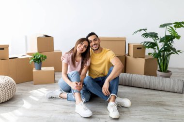 Neşeli genç bir çift yeni evlerinin zemininde oturuyor. Gülümsüyor ve rahatlıyorlar. Paketlenmemiş kutular ve yeşil bir bitkiyle çevrili. Atmosfer sıcak ve sıcak..