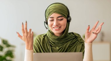 Genç bir kadın dizüstü bilgisayarın önünde otururken yeşil tesettür ve kulaklık takıyor. Gülümsüyor ve hareketli bir şekilde konuşuyor. Parlak bir ev ortamında tartışmanın tadını çıkarıyor..