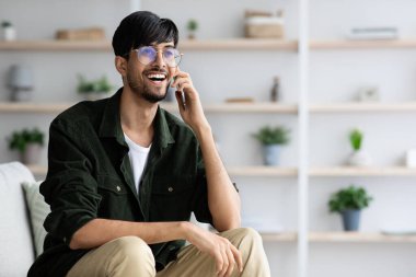 Genç bir adam kanepede oturuyor, gülümsüyor ve telefonda konuşuyor. Parlak yaşam alanı çeşitli bitkilerle dolu raflara sahip, rahat bir atmosfer yaratıyor..