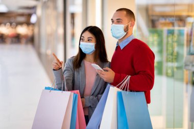 İki kişi modern bir alışveriş merkezinde alışveriş gününün tadını çıkarıyor. Maske takıyorlar ve renkli alışveriş torbaları tutuyorlar. Kadın bir mağazayı işaret ederken, adam telefonuna bakıyor..