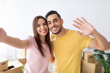 Neşeli bir çift yeni yaşam alanlarında taşınan kutularla dolu bir selfie için poz veriyor. Gülümseyip el sallıyorlar, yeni başlangıçlarını kutluyorlar..