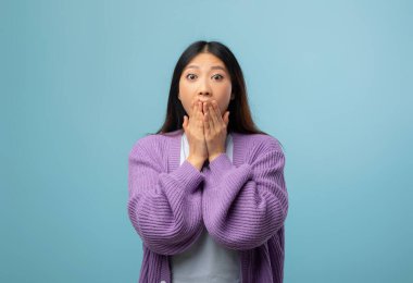 Bir kadın açık mavi bir arka planın önünde duruyor, geniş gözleri ve elleri ile ağzını kapatarak şaşkınlığını ifade ediyor. Onun uzun saçı ve mor kazağı dikkat çekici bir görünüme sahiptir..