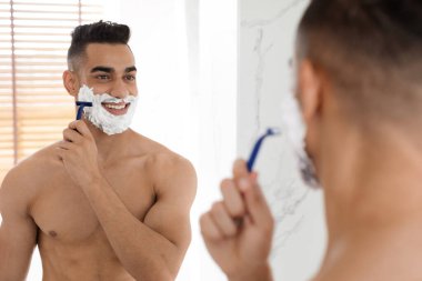 Genç bir adam banyoda sabah bakım rutininin tadını çıkarıyor. Dikkatlice tıraş kremi sürüyor ve aynadaki yansımasına bakarken sakalını tıraş etmek için tıraş bıçağı kullanıyor..