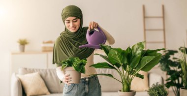 Yeşil başörtülü genç bir kadın çiçeğini parlak bir oturma odasında suluyor. Etrafı dekoratif eşyalar ve doğal ışıklarla çevrili yeşillikle ilgilenirken gülümsüyor..