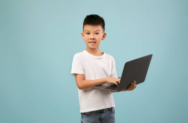 Bir çocuk elinde dizüstü bilgisayarla mutlu bir şekilde gülümsüyor. Basit bir beyaz tişört ve kot pantolon giyiyor. Açık mavi arka plan, onun neşeli ifadesini ve teknolojiye olan ilgisini vurguluyor..