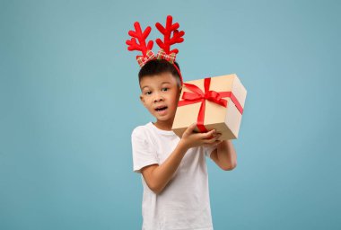 Ren geyiği boynuzu takan neşeli bir çocuk hediye paketi ile poz veriyor. Dekorun açık mavi bir arka planı var. Neşeli ve şen bir atmosfere ekleniyor..