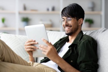 Genç bir adam kanepede rahatça oturur ve tablette okurken gülümser. Doğal ışık odayı doldurur, sıcak ve davetkar bir atmosfer yaratır..