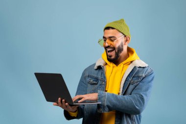 Kot ceket giyen, sarı kapüşonlu ve yeşil bereli bir adam dizüstü bilgisayarını kullanarak açık mavi bir arkaplana karşı gülümsemeyi seviyor. Çalışırken meşgul ve mutlu görünüyor..