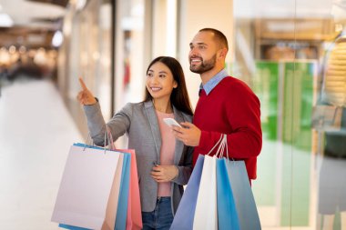 Bir erkek ve bir kadın alışveriş merkezinde alışveriş yaparken gülümserler. Renkli alışveriş torbaları tutuyorlar ve ilginç bir şeye bakıyorlar. Atmosfer canlı ve neşeli..