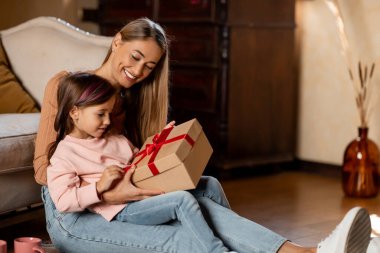 Sıcak bir oturma odasında, bir anne kızıyla oturur, kızıyla birlikte güzel paketlenmiş bir hediyeyi heyecanla açar. Gülümsemeleri, birlikteliği ve mutluluğu paylaşıyorlar..