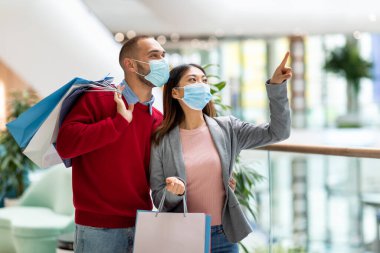 Alışveriş torbalı bir çift modern bir alışveriş merkezinin içinde duruyor. Her ikisi de yüz maskesi takıyor ve heyecanlı bir şekilde yukarı bakıyor, muhtemelen etraflarındaki mağazaları tartışıyorlar..