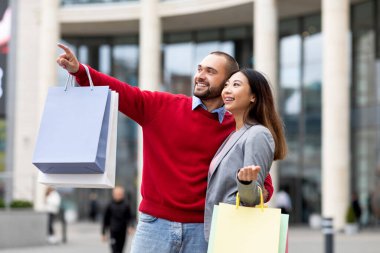 Genç bir çift şık bir alışveriş merkezinin önünde duruyor, gülümsüyor ve alışveriş torbaları tutuyorlar. İlginç bir şeye işaret ettikleri için mutlu görünüyorlar. Birlikte geçirdikleri günün tadını çıkarıyorlar..