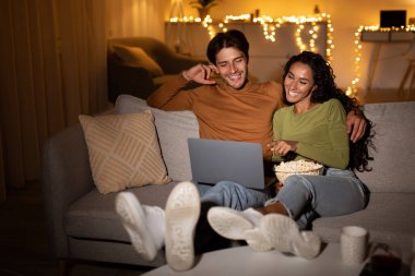 Çift rahat bir koltukta oturup dizüstü bilgisayarlarında komik bir film izliyor. Patlamış mısır ve gülümsemeleri paylaşıyorlar, sıcak ışıklarla evde rahatlatıcı bir hafta sonu akşamının tadını çıkarıyorlar..