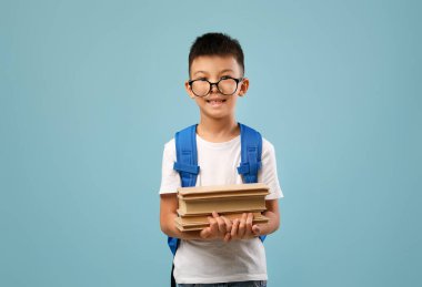 Elinde bir yığın kitap tutarken sırtüstü gülümseyen gözlüklü bir çocuk. Arka plan yumuşak bir mavidir, neşeli bir okul ortamı yaratır..