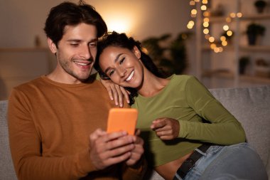 Evde rahat bir koltukta oturan çift akıllı telefon kullanmaktan mutluluk duyuyor. Kadın, kocasına gülümseyen ekranı işaret ediyor. Sahne, oturma odasındaki sıcacık ışıkla dolu..