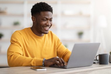 Başarılı bir siyahi adam proje yöneticisi ofiste çalışma masasında oturuyor, dizüstü bilgisayarda klavye çalıyor ve gülümsüyor, müşterilerle sohbet ediyor, işinden zevk alıyor, kopyalama alanından keyif alıyor. Evden çalışan serbest çalışan biri.