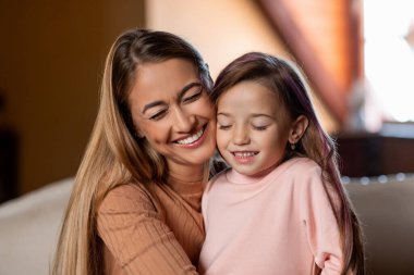 Bir anne ve küçük kızı, doğal ışıkla dolu oturma odalarında sıcacık kucaklaşırken mutlu bir şekilde gülümsüyorlar. Aralarındaki bağ neşe ve sevgi saçıyor..