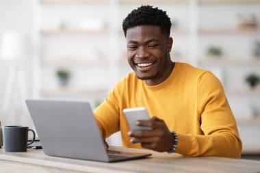 Ofiste rahat rahat oturan, dizüstü bilgisayarda çalışan ve cep telefonu kullanan neşeli genç siyahi girişimci iş için en yeni mobil uygulamaları, kopyalama alanını kontrol ediyor. Teknoloji ve iş dünyası