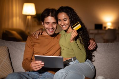 İki eş bir kanepede oturmuş, bir tablet ve bir kredi kartı kullanarak internetten alışveriş yapıyorlar. Ilık ışık, birlikte göz attıklarında sıcak bir akşam atmosferi yaratıyor..