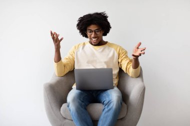 Neşeli genç siyahi adam evdeki koltukta oturuyor ve bilgisayarında video görüşmesi yapıyor. Heyecanlı görünüyor, ekrandaki biriyle konuşurken ellerini kaldırıyor..