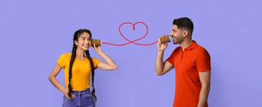 İki genç yetişkin kırmızı bir iple bağlanmış bardakları kullanarak neşeli bir anı paylaşıyorlar. Eğlenirken gülümserler. Arka plan açık mor renkli, neşeli atmosferi güçlendiriyor..