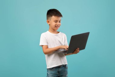 Kısa siyah saçlı bir çocuk ayakta durup dizüstü bilgisayarda yazarken gülümsüyor. Basit beyaz bir t-shirt ve kot pantolon giyiyor. Cihazla tamamen meşgul..