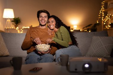 Mutlu çift oturma odasındaki kanepelerinde oturur ve duvara yansıtılan bir filmi izlerler. Bir kase patlamış mısırı paylaşıyorlar ve sıcak bir akşamın tadını çıkarıyorlar..