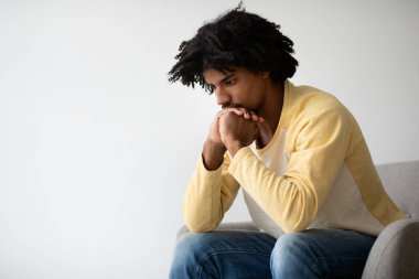 Genç siyahi bir adam evdeki koltukta düşünceli bir şekilde oturur ve çenesini ellerine yaslar. Sakin bir atmosferde sorunlarla ve zorluklarla boğuşurken düşünceli görünüyor..