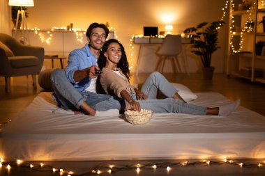 Milenyum çifti rahat bir yatakta televizyon izleyip kanal değiştiriyor. Onlar bir hafta sonu akşamında patlamış mısırın tadını çıkarıyorlar. Oda dekoratif ışıklarla hafifçe aydınlatılmış..