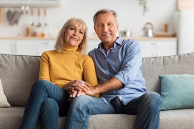 Neşeli bir çift açık gri bir kanepede oturup sıcak dekor ve doğal ışıkla dolu parlak bir oturma odasında birbirlerine eşlik ederler. Gülümsüyorlar ve el ele tutuşuyorlar..