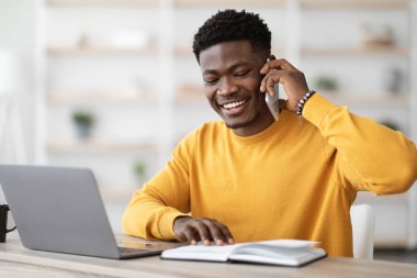 Sarı kazaklı mutlu bir zenci iş arıyor, çalışma masasında dizüstü bilgisayarla oturuyor, not defterine bakıyor, telefon görüşmeleri yapıyor ve gülümsüyor, İK ile konuşuyor, kopyalama alanı, ev içi