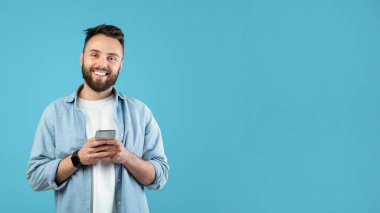 Sakallı genç bir adam gülümsüyor ve elinde bir akıllı telefon tutuyor. Sıradan bir gömlek giyiyor ve neşeli görünüyor. Parlak mavi arka plan canlı bir his katıyor..