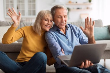 Mutlu çift, rahat bir şekilde oturma odalarında yerde otururken bir dizüstü bilgisayar görüntüsü aracılığıyla iletişim kuruyor. Gülümser ve ekranda el sallarlar, anın tadını çıkarırlar..