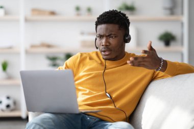 Evdeki kanepede oturan, kız arkadaşıyla görüntülü konuşma yapan, dizüstü bilgisayar ve kulaklık kullanan, defter ekranına bakıp el kol hareketi yapan, biriyle kavga eden öfkeli siyah adam.