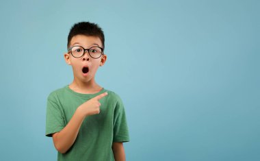 Gözlük takan genç bir çocuk ağzı açık ve parmağını göstererek hayret dolu bir bakış sergiliyor. Açık mavi arka planda yeşil bir gömlek giyiyor..